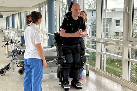 Patient im robotischen Exoskelett beim Gehtraining, begleitet von zwei Therapeutinnen im Klinikflur.