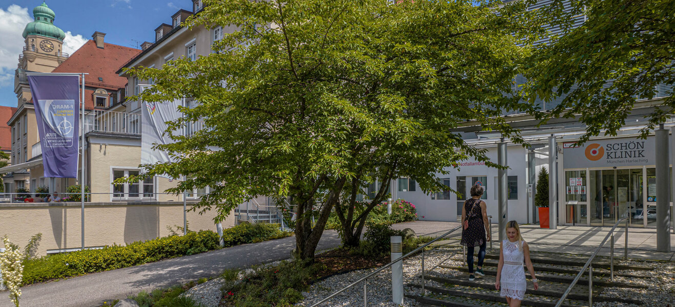 Eingangsbereich der Schön Klinik München Harlaching mit Bäumen und Besucherinnen im Sommer