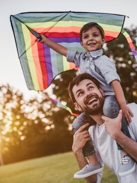 Lachender Vater trägt Kind auf den Schultern beim Drachensteigen im Sonnenschein auf einer grünen Wiese