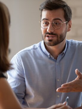 Junger Mann mit Bart und Brille spricht im modernen Büro mit einer Frau, beide am Tisch, helle Wand und Pflanze im Hintergrund