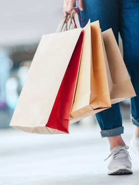 Untere Körperhälfte einer Person in blauer Jeans und grauen Sneakern, die mehrere Papiertragetaschen in der rechten Hand hält