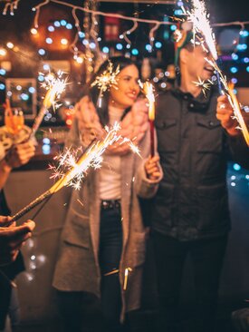 Junge Erwachsene feiern abends im Freien, halten Wunderkerzen, lachen und tragen Winterkleidung