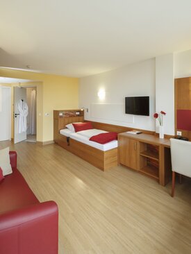 Modernes, helles Einzelzimmer mit Bett, Sessel, Schreibtisch, Fernseher und großem Fenster
