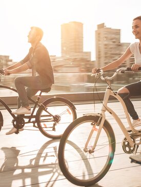 Zwei junge Erwachsene fahren bei Sonnenschein auf Fahrrädern durch eine moderne Stadt, genießen den Tag und die urbane Landschaft