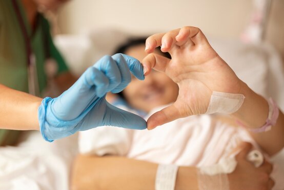 Zwei Hände, eine mit blauem Handschuh, formen ein Herz vor einer liegenden Person im Krankenhaus