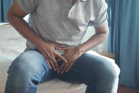 Mann sitzt auf dem Bett und hält sich mit beiden Händen den schmerzenden Unterbauch