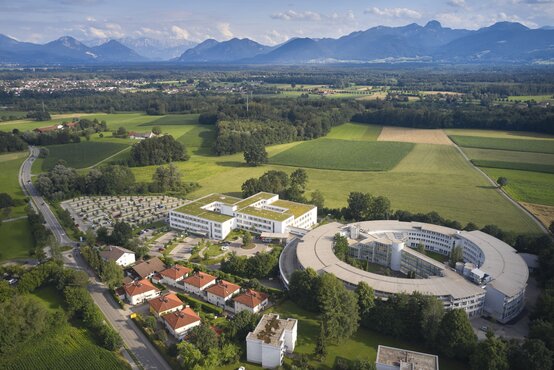 Luftaufnahme der Schön Klinik Bad Aibling Harthausen mit rechteckigem und rundem Bau, umgeben von Grünflächen und Bergen im Hintergrund