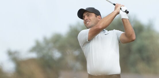 Männlicher Golfer im weißen Poloshirt und dunkler Kappe schlägt mit Golfschläger einen Ball auf dem Golfplatz