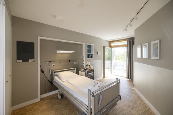 Modernes, helles Patientenzimmer mit Einzelbett, Nachttisch, Flachbildfernseher, Balkonzugang, Holzfußboden und viel Tageslicht