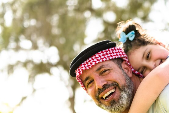 Lächelnder arabischer Vater mit rot-weißer Kufiya trägt seine Tochter mit blauer Schleife draußen an einem sonnigen Tag
