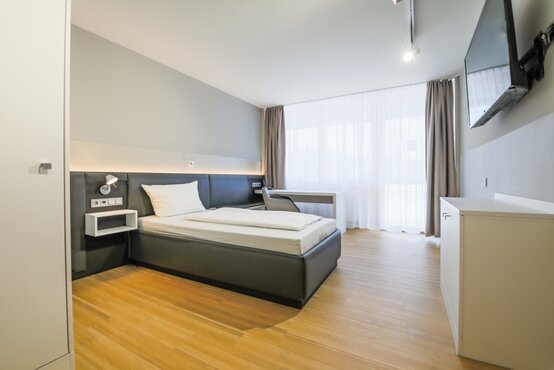 Modernes Einzelzimmer mit großem Fenster, Einzelbett, Schreibtisch, TV und heller, komfortabler Einrichtung