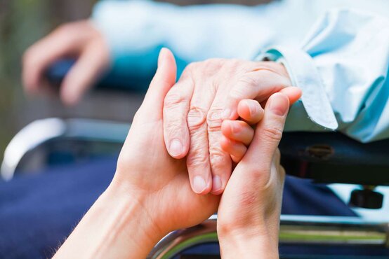 Junge Hand hält unterstützend die Hand einer älteren Person auf einem Rollstuhl