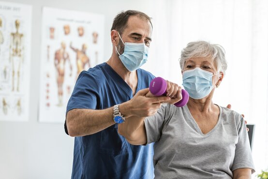 Physiotherapeut betreut ältere Patientin bei Übung mit Hanteln in Klinik, beide mit Maske, Fokus auf Rehabilitation