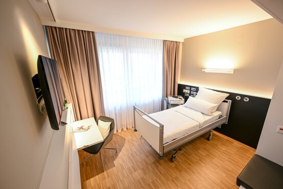 Einzelzimmer in der Klinik mit Krankenbett, Nachttisch, Schreibtisch, Fernseher und viel Tageslicht