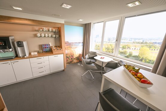 Moderne, helle Klinik-Lounge mit Fensterfront, Sitzgruppen, Stehtisch mit Obstschale und Kaffee-Getränkestation