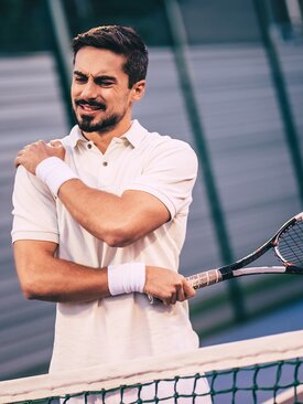 Junger Mann auf Tennisplatz greift sich mit schmerzverzerrtem Gesicht an die Schulter, Tennisschläger in der anderen Hand
