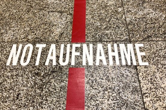 Schriftzug „Notaufnahme“ in weißen Großbuchstaben auf grau-gesprenkeltem Boden, breite rote Linie verläuft senkrecht durch das Wort