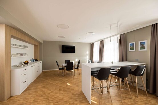 Moderner, heller Loungebereich mit weißer Anrichte, Barhockern, Obstkorb, Tischen, schwarzen Stühlen und großem Fenster