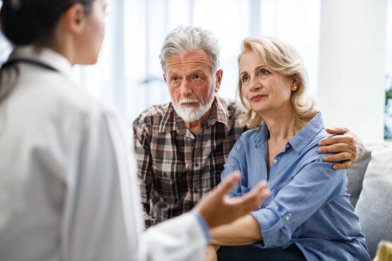 Ärztin berät älteres Paar auf Sofa in heller Klinik, Mann legt schützend Arm um Frau.