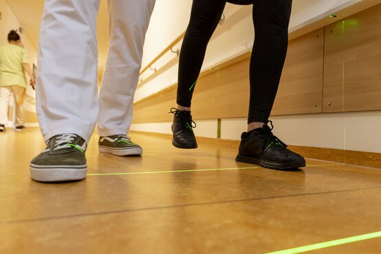 Zwei Personen gehen im Klinikflur über grüne Laserlinien zur Gangschulung, Handlauf an der Wand