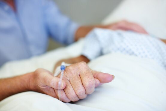 Ältere Patientin mit Infusion im Krankenhausbett, deren Hand von einer zweiten Person fürsorglich gehalten wird