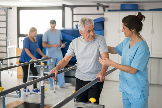 Physiotherapeutin unterstützt älteren Mann beim Gehen an Parallelbarren in heller Therapiepraxis