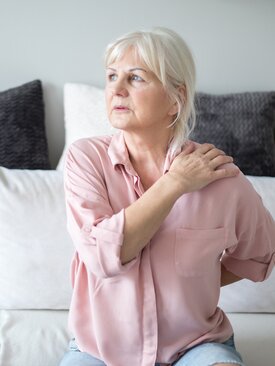 Ältere Frau sitzt auf einem Sofa und hält sich die linke Schulter