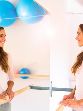 Junge Frau an Ballettstange im hellen Fitnessraum, lächelt in Spiegel und ist zufrieden