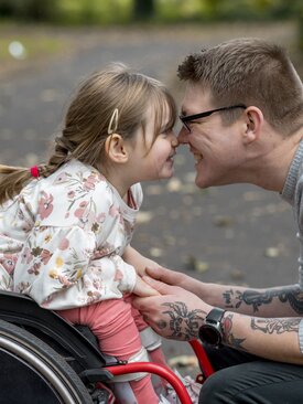 Erwachsener mit Brille und Tattoos hält die Hände eines Mädchens im Rollstuhl, beide lächeln sich an und berühren die Nasen