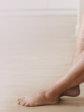 Unterschenkel und Füße einer sitzenden Person mit sichtbaren Venen und Hautunebenheiten, Fokus auf Venengesundheit.