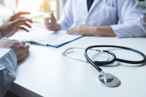 Stethoskop liegt auf einem weißen Tisch, im Hintergrund führen zwei Personen ein Gespräch, eine davon trägt einen weißen Arztkittel