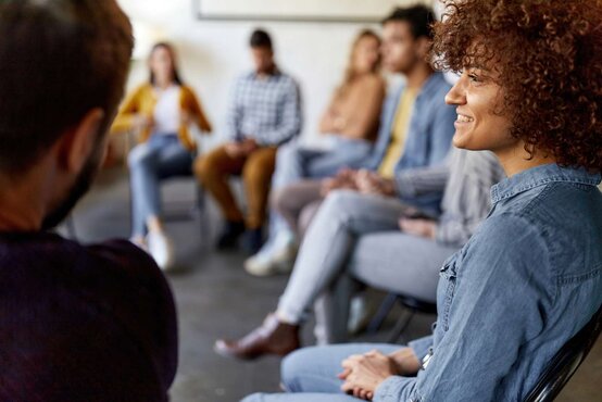 Junge Frau im Jeanshemd lächelt im Stuhlkreis während Gruppentherapie, andere Teilnehmer sitzen unscharf im Hintergrund