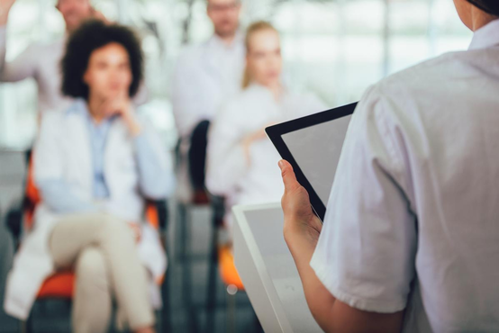 Vortragende Person im weißen Kittel präsentiert mit Tablet vor Medizinerteam bei Präsentation im modernen Seminarraum