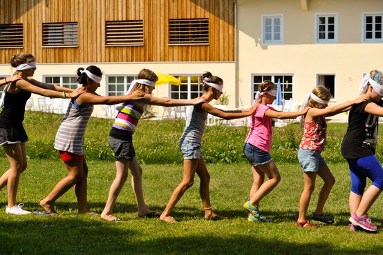 Kinder laufen mit verbundenen Augen und Händen auf den Schultern des Vordermanns über grüne Wiese