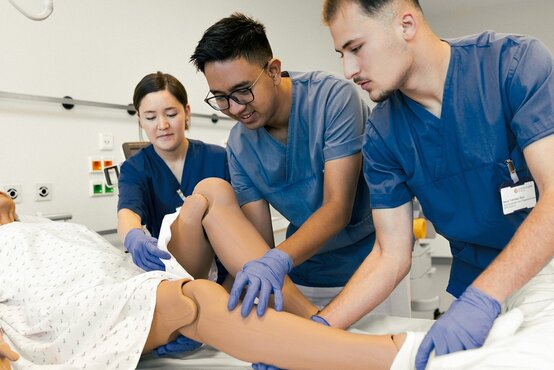 Drei Pflegekräfte üben gemeinsam Pflegetechniken an Pflegepuppe im modernen Krankenhauszimmer