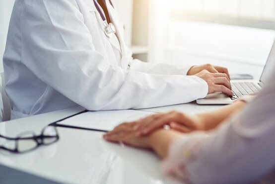 Arzt im weißen Kittel mit Stethoskop tippt am Schreibtisch auf Laptop, Patientin sitzt gegenüber