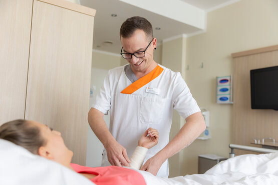 Pflegekraft in weißer Kleidung versorgt Arm einer Patientin mit Verband im modernen Krankenhauszimmer.