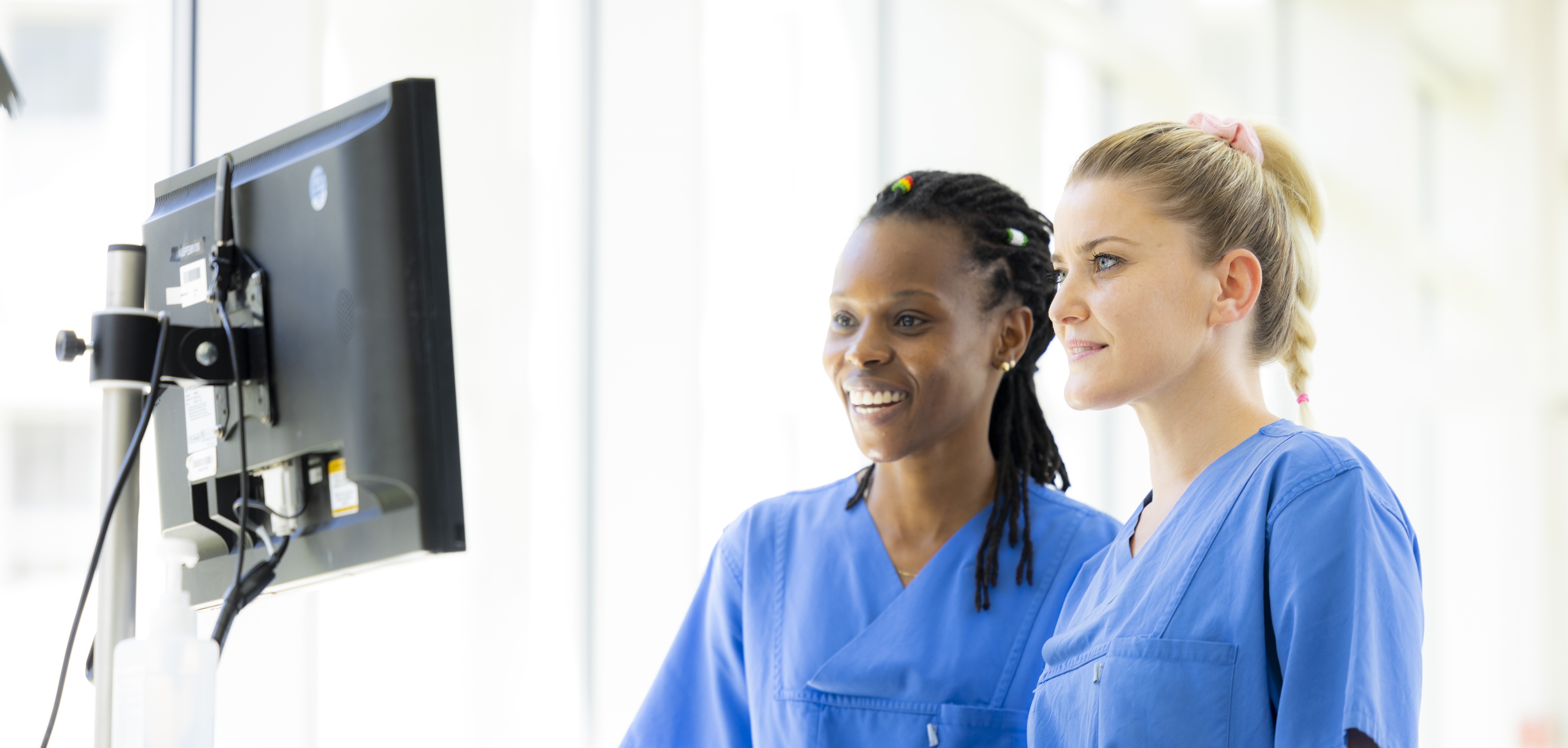 Zwei Pflegekräfte in blauer Kleidung betrachten gemeinsam Monitor im hellen Klinikraum, Teamarbeit.