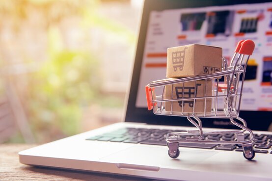 Kleiner Einkaufswagen mit Kartons auf Laptop-Tastatur, unscharfer Bildschirm mit Online-Shop im Hintergrund