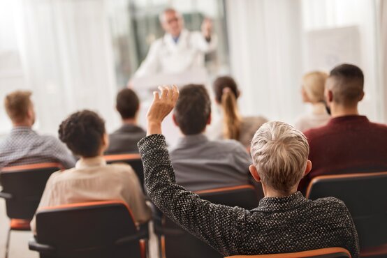 Arzt hält Vortrag vor Zuhörergruppe, Personen sitzen mit dem Rücken zur Kamera.