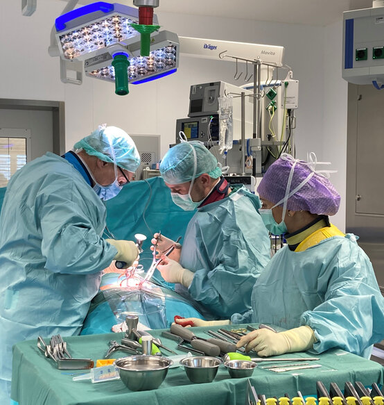 Drei Chirurgen in steriler OP-Kleidung operieren gemeinsam einen Patienten im modernen Operationssaal mit chirurgischen Instrumenten