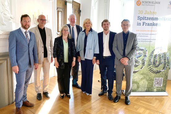 Sieben Gratulanten in festlicher Kleidung vor Jubiläumsbanner „30 Jahre Schön Klinik Bad Staffelstein“ auf Kloster Banz