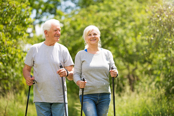 Älteres Paar geht lächelnd auf sonnigem Naturweg beim Nordic Walking mit Stöcken, Bewegungstherapie im Grünen