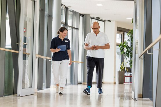 Therapeutin begleitet Parkinson-Patient im modernen Klinikflur mit Handläufen und Tageslicht.