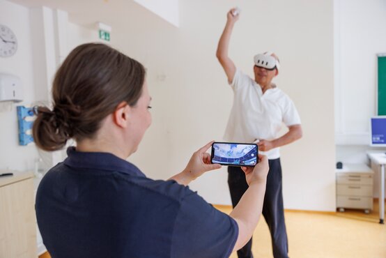 Person mit VR-Brille und Ball macht Bewegungstraining, zweite Person überwacht per Smartphone.