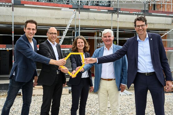 Fünf Personen in Businesskleidung halten gemeinsames gelbes Symbol und lächeln vor Baustelle des Ronald McDonald Hauses Vogtareuth