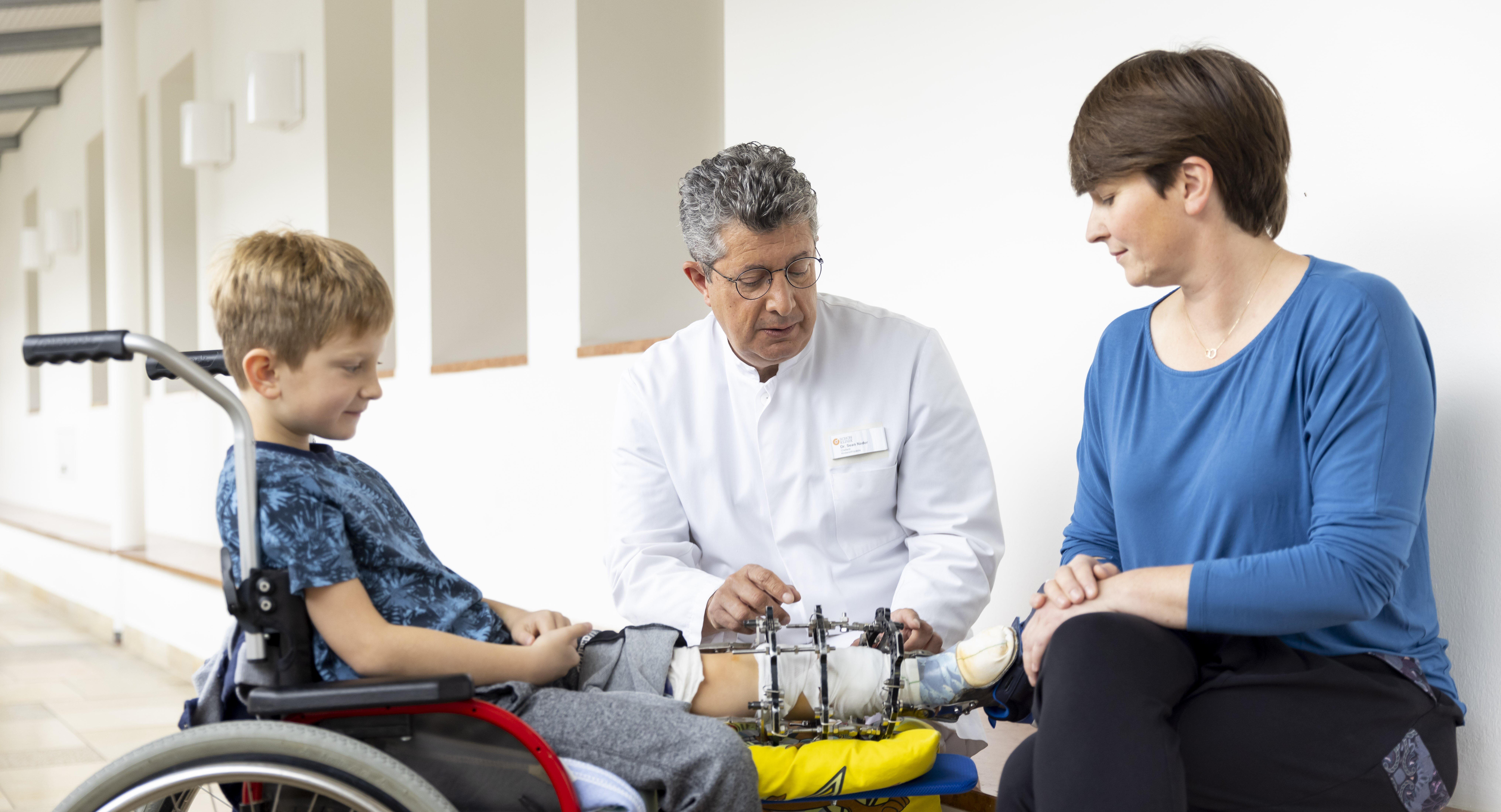 Arzt im weißen Kittel erklärt Jungen im Rollstuhl mit äußerem Fixateur am Bein und Mutter das Bein in hellem, freundlichem Klinikflur