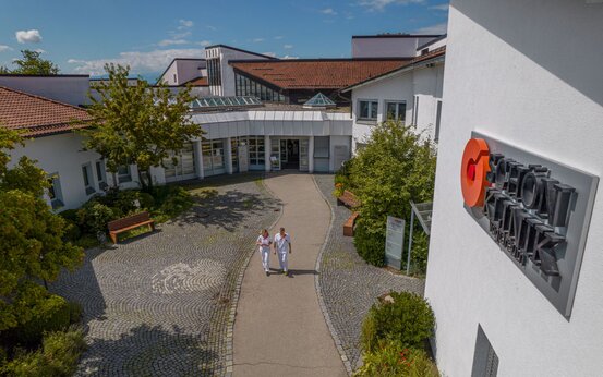 Außenansicht der Schön Klinik Vogtareuth mit rotem Dach, zentralem Eingang, zwei Personen in Weiß und Kliniklogo