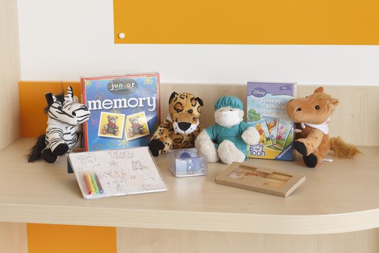 Ablage in Kinderecke mit Zebra-, Geparden- und Pferdeplüschtier, Krankenhauspuppe, Memory- und Schnipp-Schnapp-Spiel, Bastelset vor orangem Wandpaneel