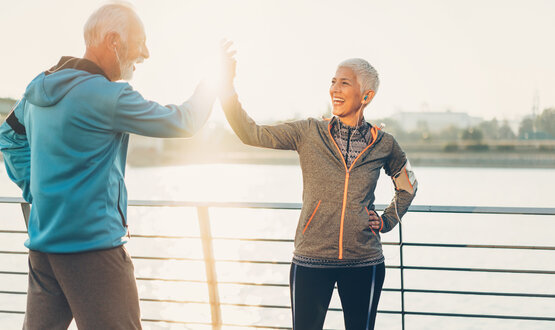 Älteres Ehepaar in Sportkleidung gibt sich lachend High-Five an Flussgeländer, Ohrhörer, Handytasche, sonniges Wetter